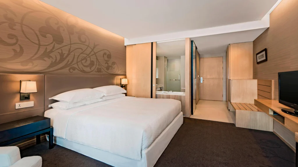 Modern hotel room with a large bed, nightstands with lamps, a TV on a wooden stand, light-colored walls with a decorative pattern, and a partial view of a bathroom through glass doors.