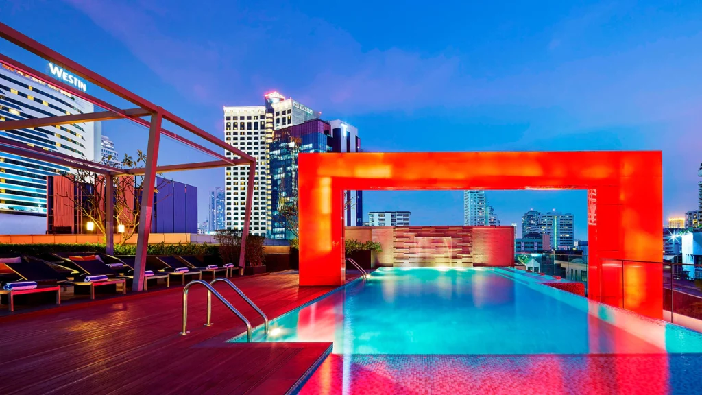 A modern rooftop pool at dusk, illuminated by vibrant red and blue lights, with city skyscrapers in the background and lounge chairs along the wooden deck.