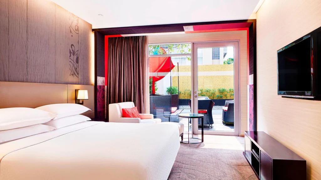 A modern hotel room with a large bed, armchair, wall-mounted TV, and a glass door opening to a furnished outdoor patio with plants and a red umbrella.