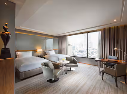 Modern hotel room with two double beds, a large window with city views, a small round table with two chairs, a desk with a lamp, and neutral-toned decor.