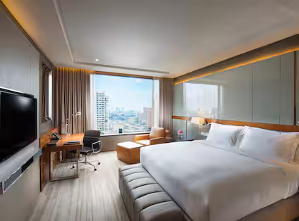Modern hotel room with a large bed, a wall-mounted TV, a desk with a chair, a lounge chair, and a large window offering a city view. The room is bright with neutral colors and minimalist decor.