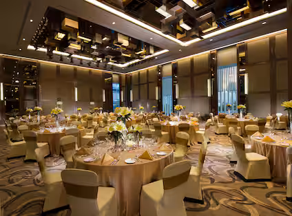 Elegant banquet hall with round tables covered in gold cloth, yellow floral centerpieces, banquet chairs, elaborate ceiling lights, and patterned carpet, set up for a formal event or reception.