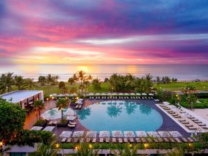 A large, round swimming pool surrounded by lounge chairs and palm trees overlooks a lush green lawn and the ocean at sunset, with a vibrant pink, purple, and orange sky.