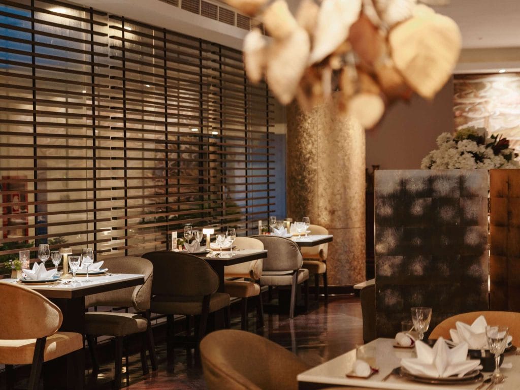 A stylish restaurant interior with neatly set tables, elegant chairs, folded napkins, glasses, and soft lighting. Decorative elements and a wooden partition create a cozy, upscale atmosphere.
