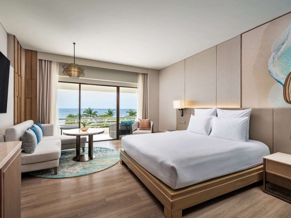 Modern hotel room with a large bed, sofa, round table, and balcony overlooking palm trees and the ocean. The room has light-colored walls, wooden floors, and a nature-inspired wall design above the bed.