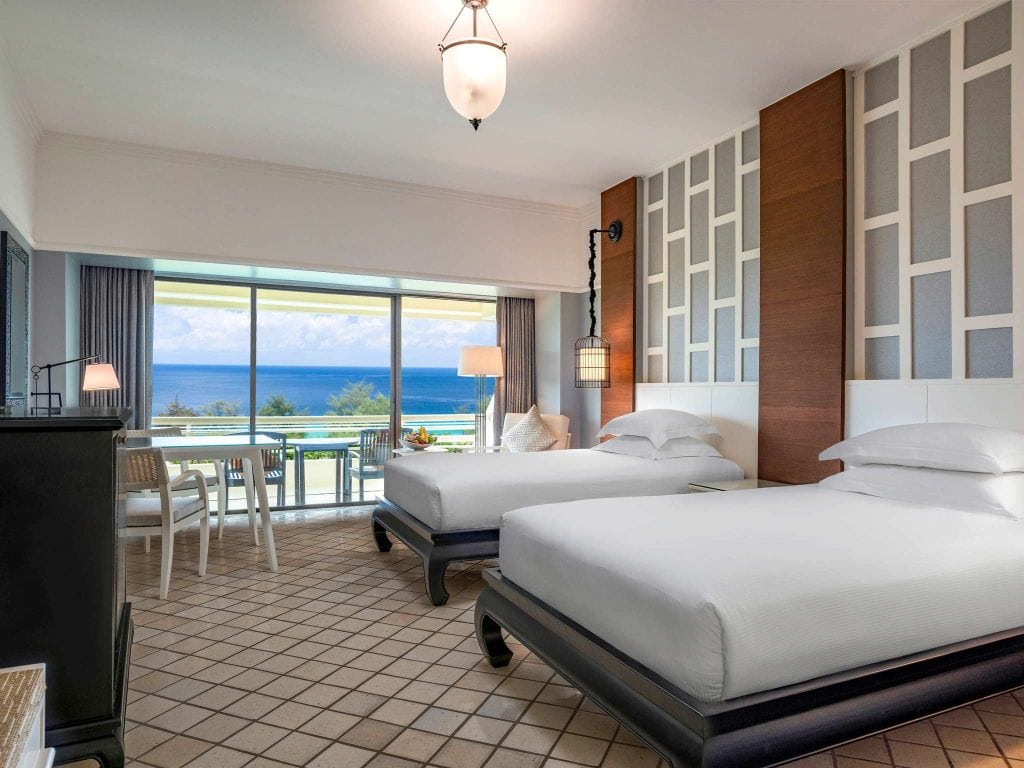 A modern hotel room with two twin beds, geometric accent wall, tiled floor, and large window opening to a balcony with a table and chairs, overlooking the ocean and blue sky.