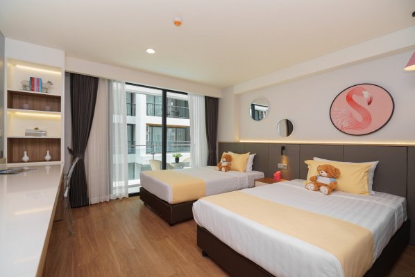 A modern hotel room with two single beds, each with yellow and white bedding and a teddy bear on the pillow. There is a desk, open shelves, a large window with curtains, and a flamingo artwork on the wall.