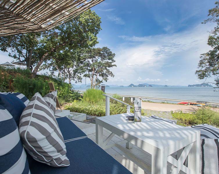 A shaded patio with blue and white striped cushions and a white table overlooks a sandy beach and calm sea, with distant islands and a blue sky in the background. Trees and greenery surround the relaxing seaside spot.