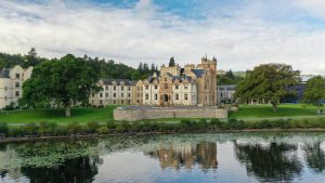 A large, historic castle-like building with a tower sits on a manicured lawn by a calm river, surrounded by trees and reflected in the water under a partly cloudy sky.