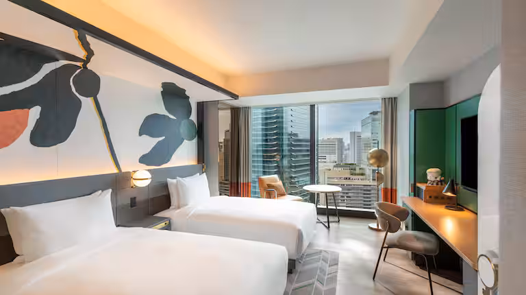 Modern hotel room with two white beds, abstract wall art, large windows with a city view, a desk, chairs, and contemporary decor in neutral and green tones.