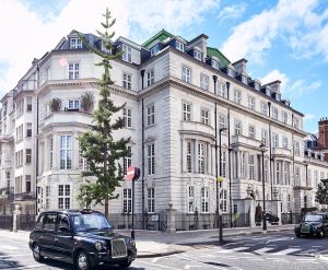 A large, elegant white stone building with many windows and decorative details on a sunny day; a black London taxi passes in front, and a tall tree stands by the corner.