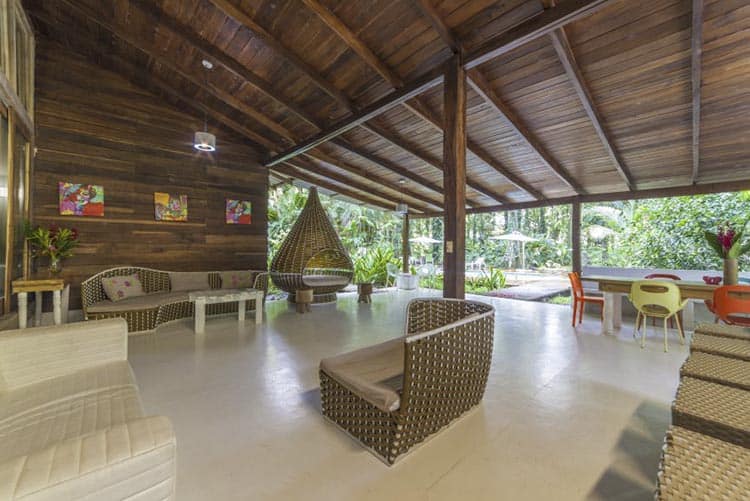 Open-air lounge area with wicker furniture, colorful chairs, and a high wooden ceiling. Large windows reveal lush greenery and a pool outside, creating a relaxed tropical atmosphere.