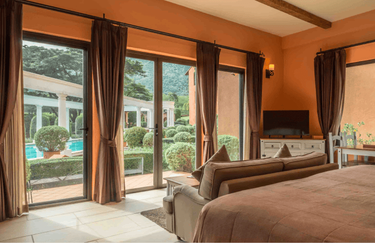 A cozy bedroom with brown decor and large glass doors overlooking a garden, manicured bushes, and a swimming pool with surrounding columns and greenery outside.