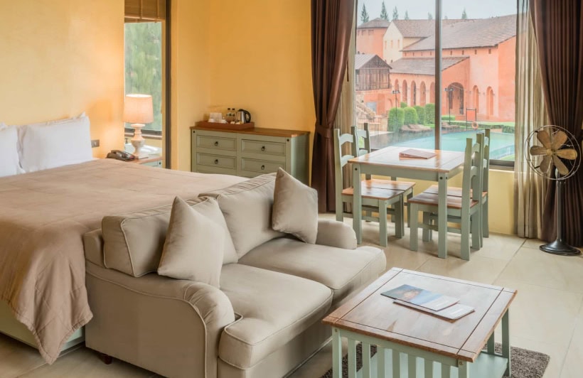 A cozy hotel room with a double bed, sofa, coffee table, dining table with four chairs, and large windows overlooking a scenic courtyard with orange buildings and greenery.