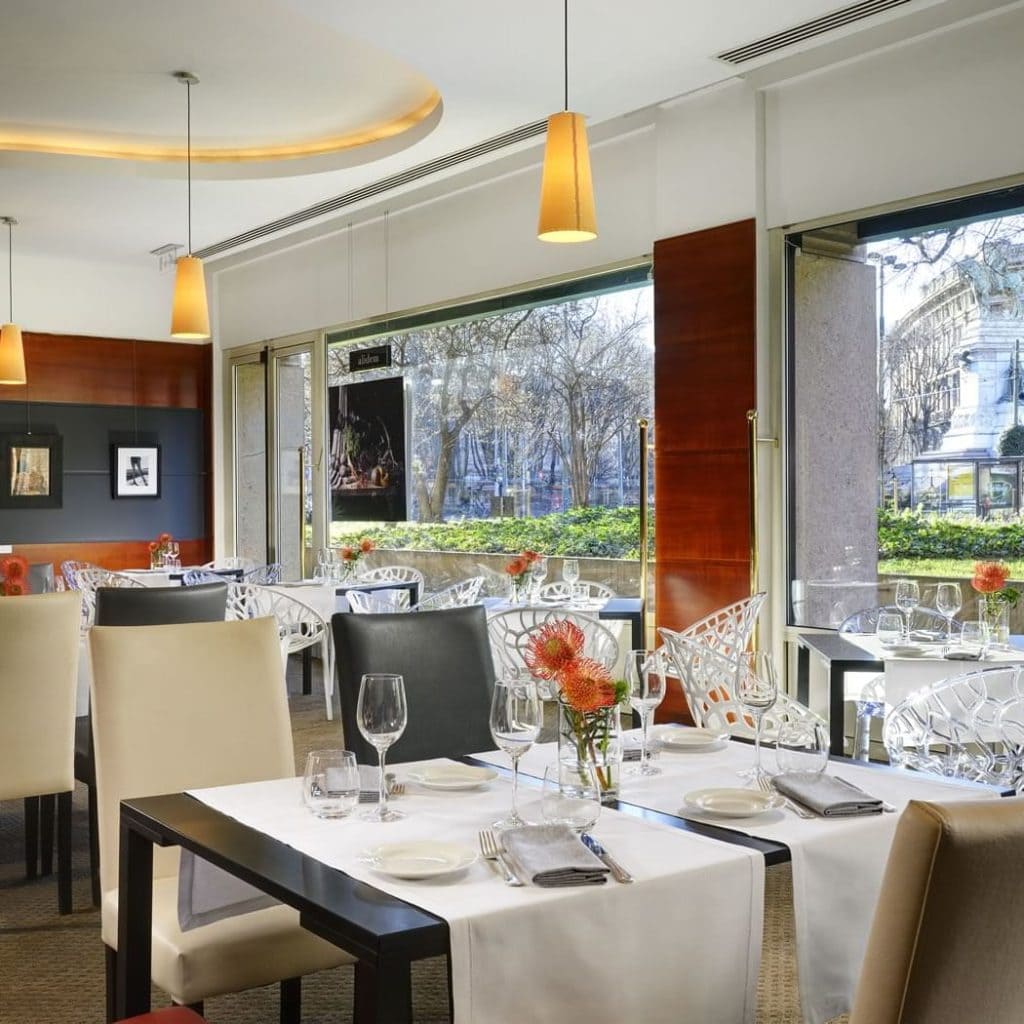 Elegant restaurant interior with neatly set tables, modern chairs, large windows offering a street view, and pendant lights. Orange flowers decorate each table, and natural light fills the inviting dining space.