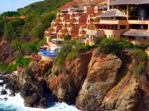 A resort with tan buildings and thatched roofs is perched on a rocky cliff above the ocean, surrounded by lush greenery and overlooking waves crashing against the rocks below.