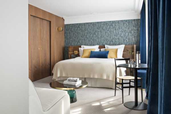 A modern hotel room with a double bed, blue and gold accent pillows, a textured blue accent wall, a round coffee table, a small dining table with two chairs, and large blue curtains.