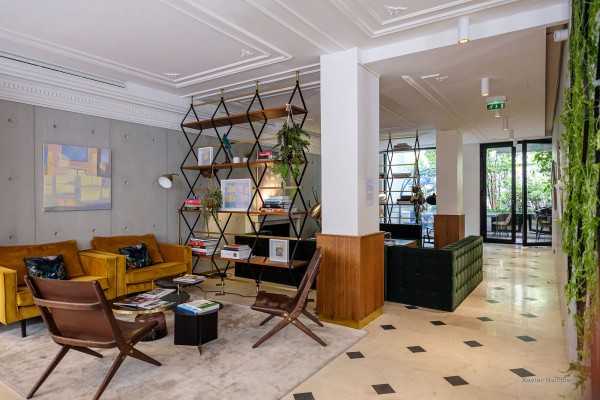 Modern lounge with mustard yellow sofas, brown chairs, a geometric bookshelf, and green plants. Large windows let in natural light; marble floors and white walls give a clean, contemporary feel.