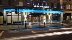 Nighttime view of the entrance to a DoubleTree by Hilton hotel, featuring illuminated blue lights above the doors and blurred light trails from passing vehicles in front of the building.