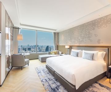 Modern hotel room with a large bed, accent pillows, armchair, ottoman, and floor-to-ceiling window offering a city skyline view. Light-colored decor and wooden flooring create a bright, inviting atmosphere.