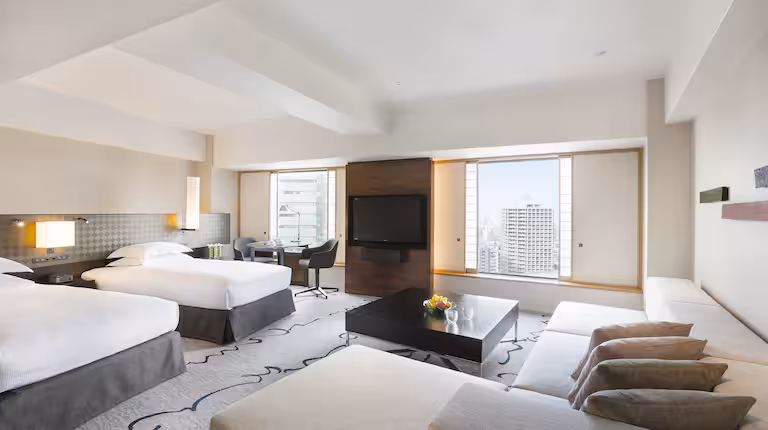 Modern hotel room with two large beds, a seating area with a coffee table and sofa, a wall-mounted TV, and large windows offering a city view. The room is bright and decorated in neutral tones.