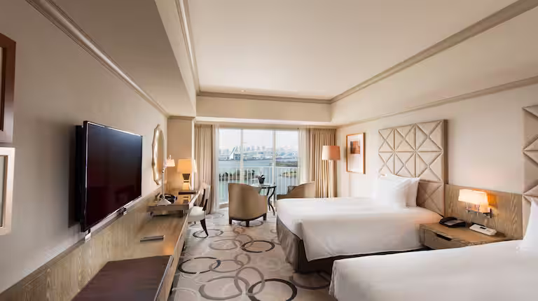 A modern hotel room with two double beds, a flat-screen TV, a desk, and a sitting area near large windows overlooking a balcony and a scenic city or waterfront view. Neutral colors and contemporary decor create a welcoming atmosphere.