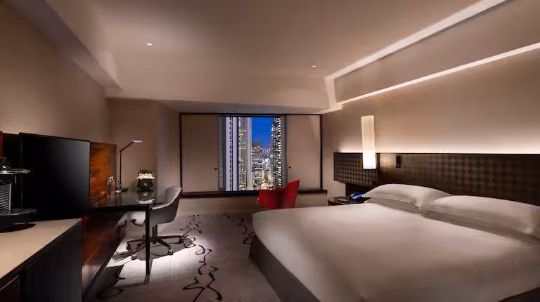 A modern hotel room with a large bed, desk, and chair. Floor-to-ceiling windows reveal a cityscape at night. The room has warm lighting and contemporary decor.