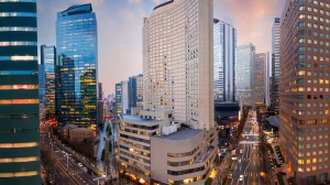 Cityscape at dusk showing tall, modern skyscrapers with glass and concrete facades, busy streets filled with cars, and city lights starting to illuminate as daylight fades.