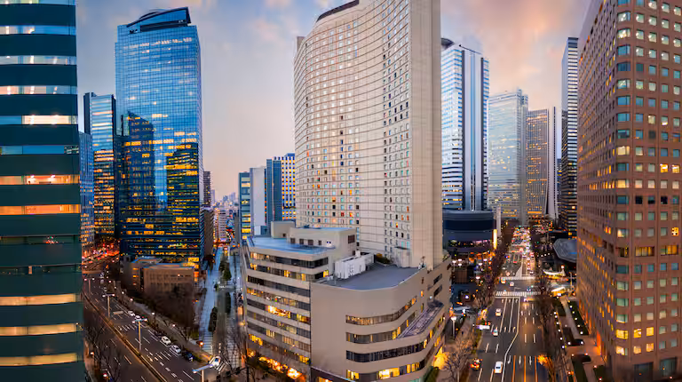Cityscape at dusk showing tall, modern skyscrapers with glass and concrete facades, busy streets filled with cars, and city lights starting to illuminate as daylight fades.