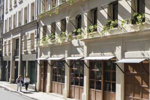 Sunlit street with two women walking past a beige building featuring large wooden windows, decorative window awnings, and greenery on the upper window ledges.