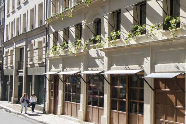 Sunlit street with two women walking past a beige building featuring large wooden windows, decorative window awnings, and greenery on the upper window ledges.