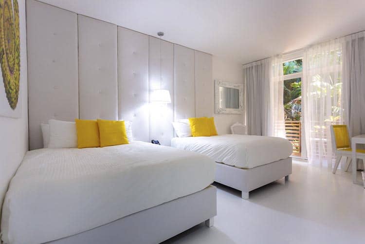 A bright hotel room with two white beds, each accented with yellow pillows, white walls and floors, sheer curtains, a small desk, a mirror, and large windows showing greenery outside.