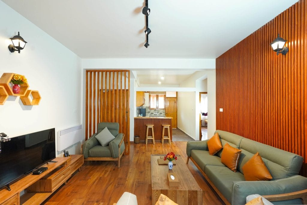 Modern living room with wooden flooring and paneling, green sofas with orange pillows, a TV on a wooden stand, armchair, decorative wall shelves, and an open view into a kitchen with bar stools.