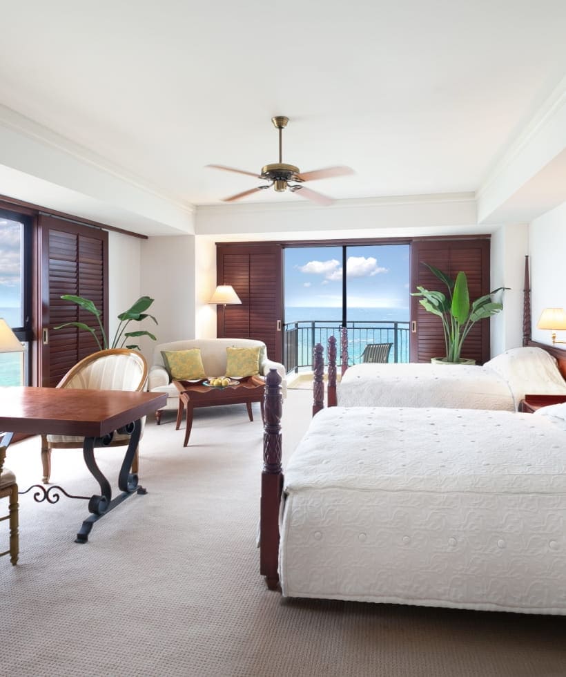 A bright hotel room with two double beds, a wooden table and chairs, a sofa, large windows with wooden shutters, potted plants, and a balcony overlooking the ocean under a blue sky.