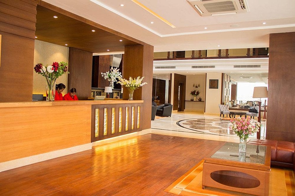A modern hotel lobby with wooden floors, a reception desk staffed by two people, floral arrangements, comfortable seating, and bright lighting. The atmosphere is welcoming and spacious.