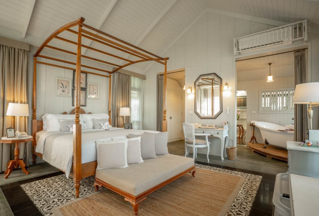 A bright, airy bedroom with a wooden four-poster bed, white linens, a cushioned bench, a writing desk with a mirror, a freestanding bathtub, and elegant décor in neutral tones. Light streams in from large windows.