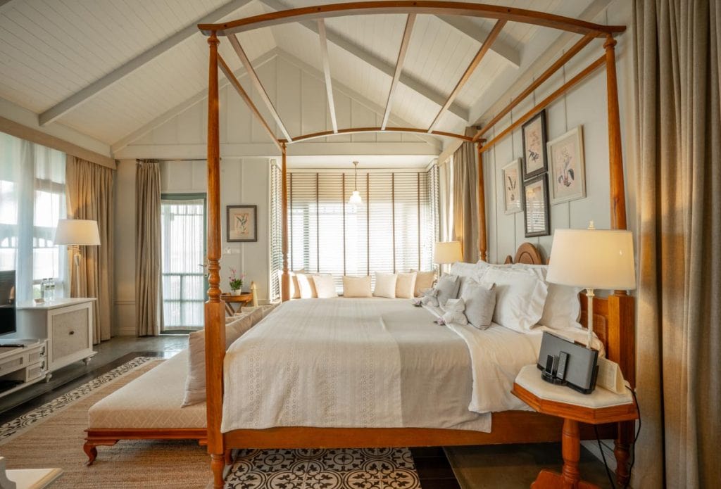A cozy bedroom with a wooden four-poster bed, white bedding, decorative pillows, bedside lamps, a bench at the foot of the bed, large windows with blinds, and soft natural light filling the room.
