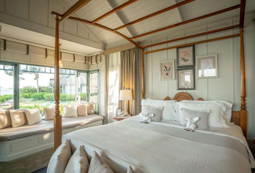 A bright, cozy bedroom with a four-poster bed, crisp white linens, and towel animals. A window seat with cushions overlooks a lush garden and palm trees outside. Soft lighting and framed art create a relaxing atmosphere.