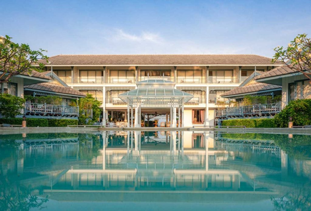 A modern resort building with balconies is reflected in a large swimming pool, surrounded by greenery and clear blue sky.