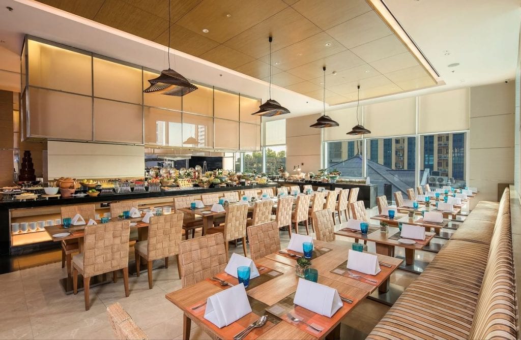 Modern restaurant dining area with wooden tables set with white napkins, blue glasses, and wicker chairs; buffet counters with assorted food are visible in the background under warm lighting. Large windows let in natural light.