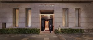 The entrance of The Celestine building at night, featuring a large wooden door, two tall windows with warm lighting, and a person in a suit standing at the doorway. Green shrubs line the front of the building.