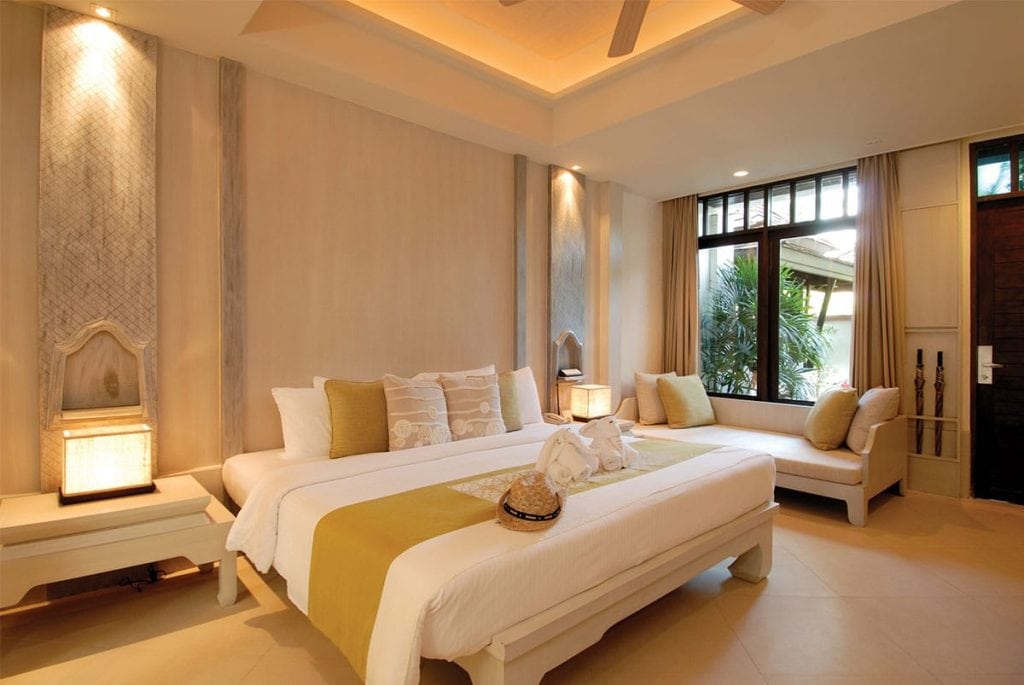 A modern, elegant bedroom with a large bed, white and beige decor, yellow accents, two swan-shaped towels on the bed, a cushioned bench by a window, and warm ambient lighting.