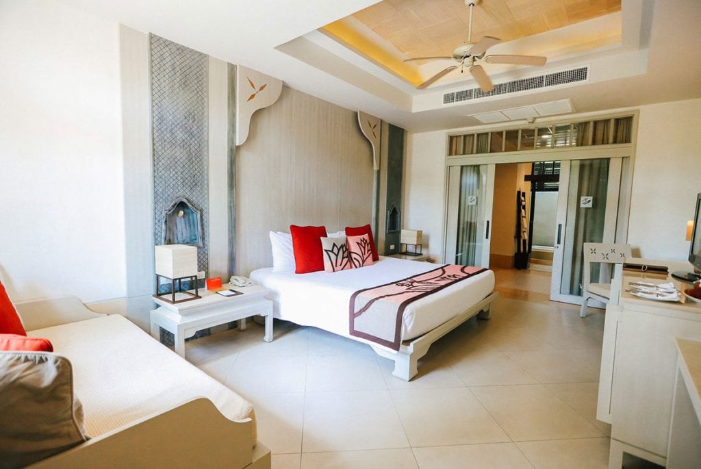 A bright, modern hotel room with a large bed, red and white pillows, two bedside tables, a ceiling fan, a sofa, and a glass wall separating the bedroom from the bathroom area.