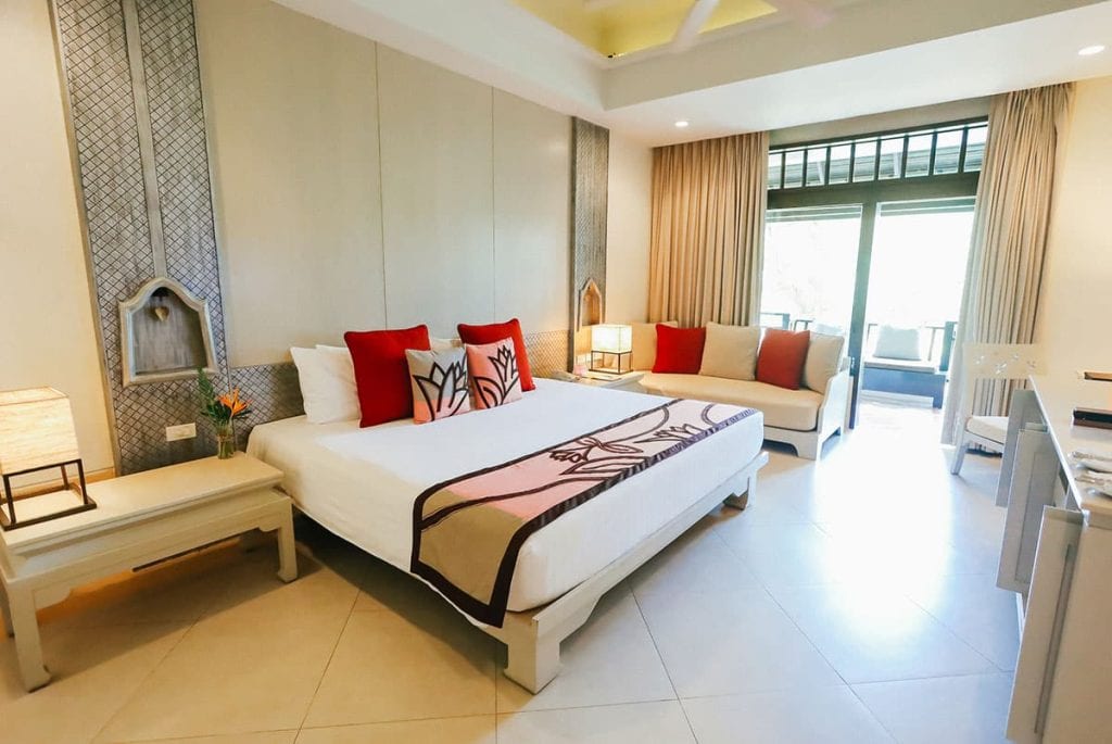Modern hotel room with a large bed featuring red and patterned pillows, a side table with a lamp, a sofa by the window, and glass doors leading to a balcony with natural light streaming in.