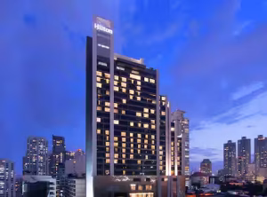 Tall Hilton hotel building with illuminated windows, set against a dusky blue sky and surrounded by other high-rise buildings in a cityscape.