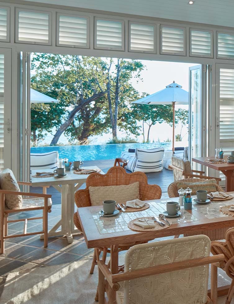 A sunlit dining area with wooden tables and chairs overlooks a pool and outdoor lounge with umbrellas and trees, set against a backdrop of blue sky and water. Breakfast settings are arranged on the tables.
