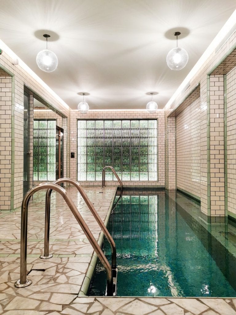 A narrow indoor swimming pool with mosaic tile walls and floor, metal handrails, and modern globe ceiling lights. A large window at the end lets in natural light, giving the space a clean, serene atmosphere.