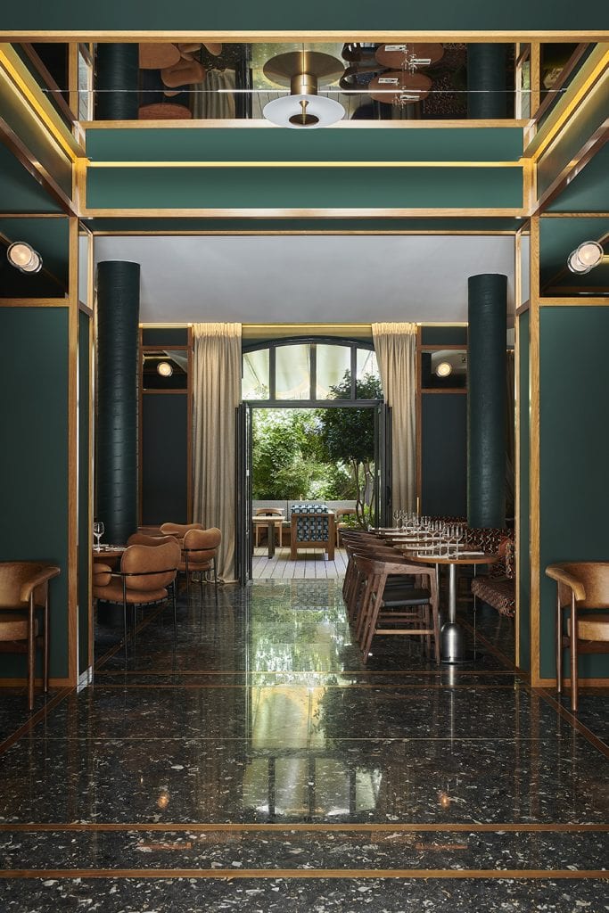 Elegant restaurant interior with dark green walls, gold accents, marble floors, brown leather chairs, wooden tables set for dining, large windows, and sheer curtains revealing a lush, green outdoor space.