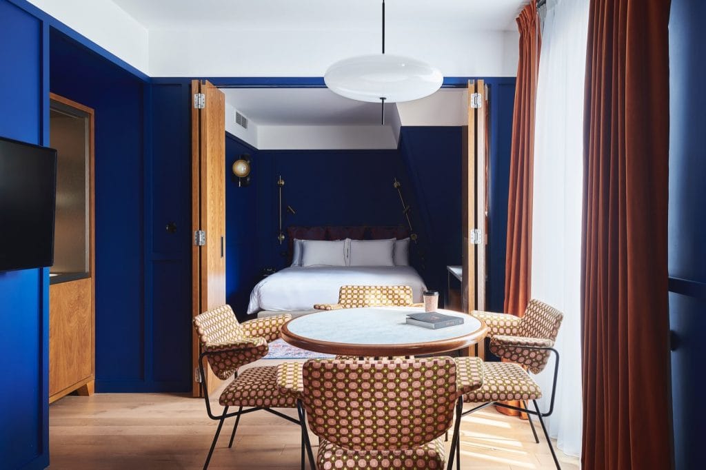 A modern hotel room with deep blue walls, a bed with white linens in a cozy alcove, a round table surrounded by patterned chairs, and large windows with burnt orange curtains letting in natural light.