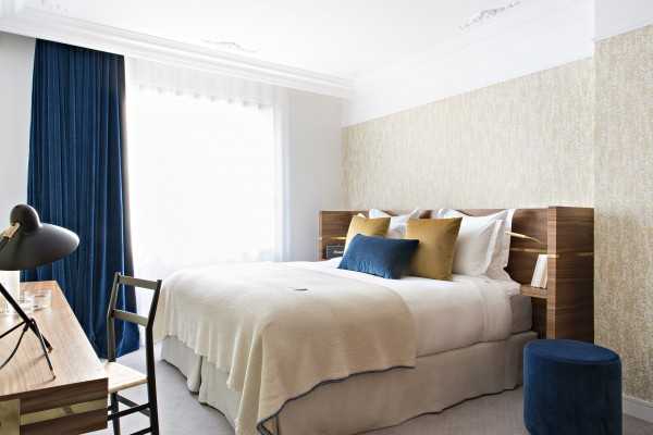Modern bedroom with a large bed featuring beige bedding, blue and yellow accent pillows, dark blue curtains, a wooden desk with a black lamp, and a blue ottoman, all set in a bright, well-lit space.